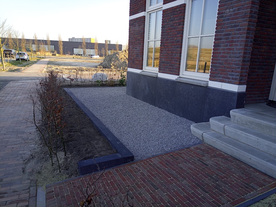Voortuin bij een nieuwbouwwoning met grind, een zwarte opsluitband, klinkers en een jonge haag
