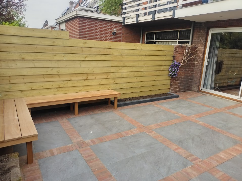 Terras met grijze tegels en rode klinkerbanden, een houten schutting en een houten hoekbank
