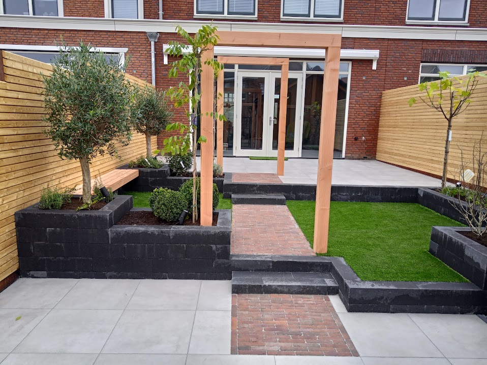 Achtertuin met terras, trap, keerwanden van zwarte blokken, kunstgras, een klinkerpad onder een houten pergola en olijfbomen