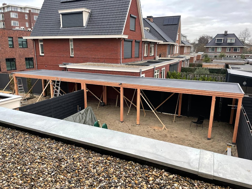 Lange houten overkapping in aanbouw achter een rij woningen, met zand in de tuin