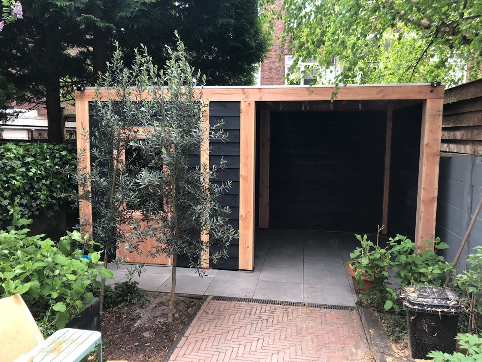 Tuinhuis met donkere houten wanden en Douglas staanders, een visgraat klinkerpad en een jong olijfboompje