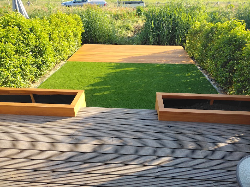 Tuin met vlonder, kunstgras, twee houten plantenbakken en hagen aan weerszijden